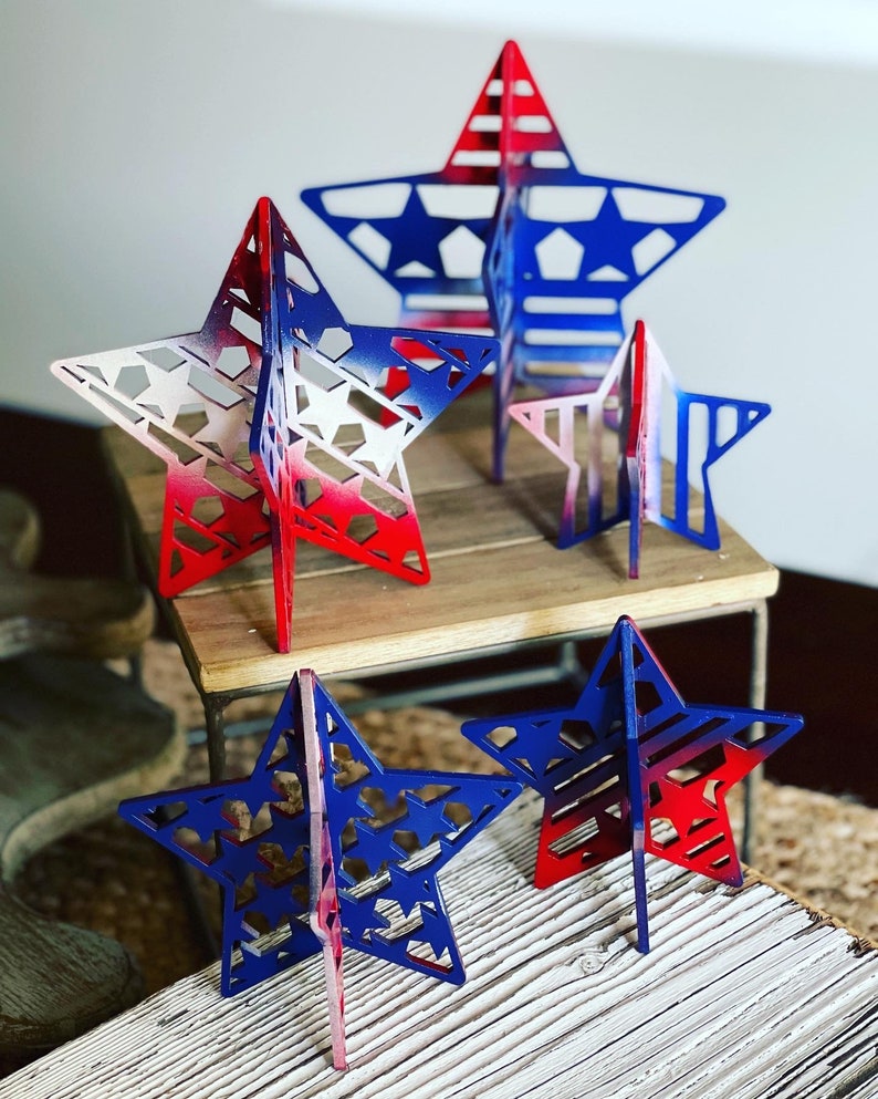 May include: Four wooden stars painted in red, white and blue with a patriotic theme. The stars are different sizes and are standing on a wooden surface.