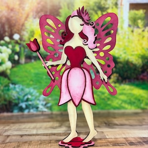 May include: A wooden fairy figurine with pink and red wings, a pink dress, and a flower wand. The fairy is standing on a pink base.