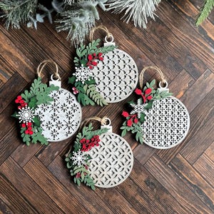 May include: Four white wooden Christmas ornaments with a geometric pattern. Each ornament is decorated with green leaves, red berries, and a white snowflake.
