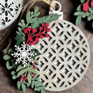 May include: A white wooden Christmas ornament with a geometric pattern and a green and red floral design. The ornament is hanging from a twine string.