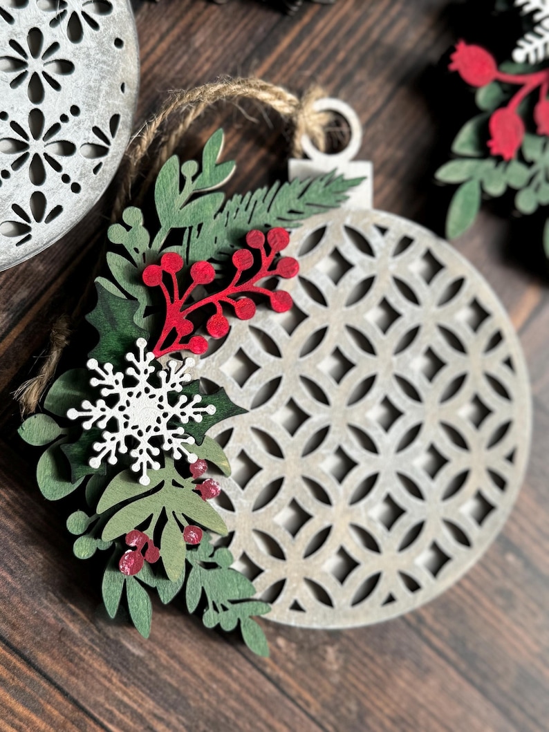 May include: A white wooden Christmas ornament with a geometric pattern and a green and red floral design. The ornament is hanging from a twine string.