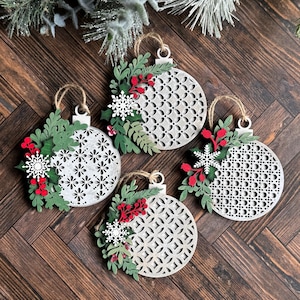 May include: Four white wooden Christmas ornaments with a geometric pattern. Each ornament is decorated with green leaves, red berries, and white snowflakes.