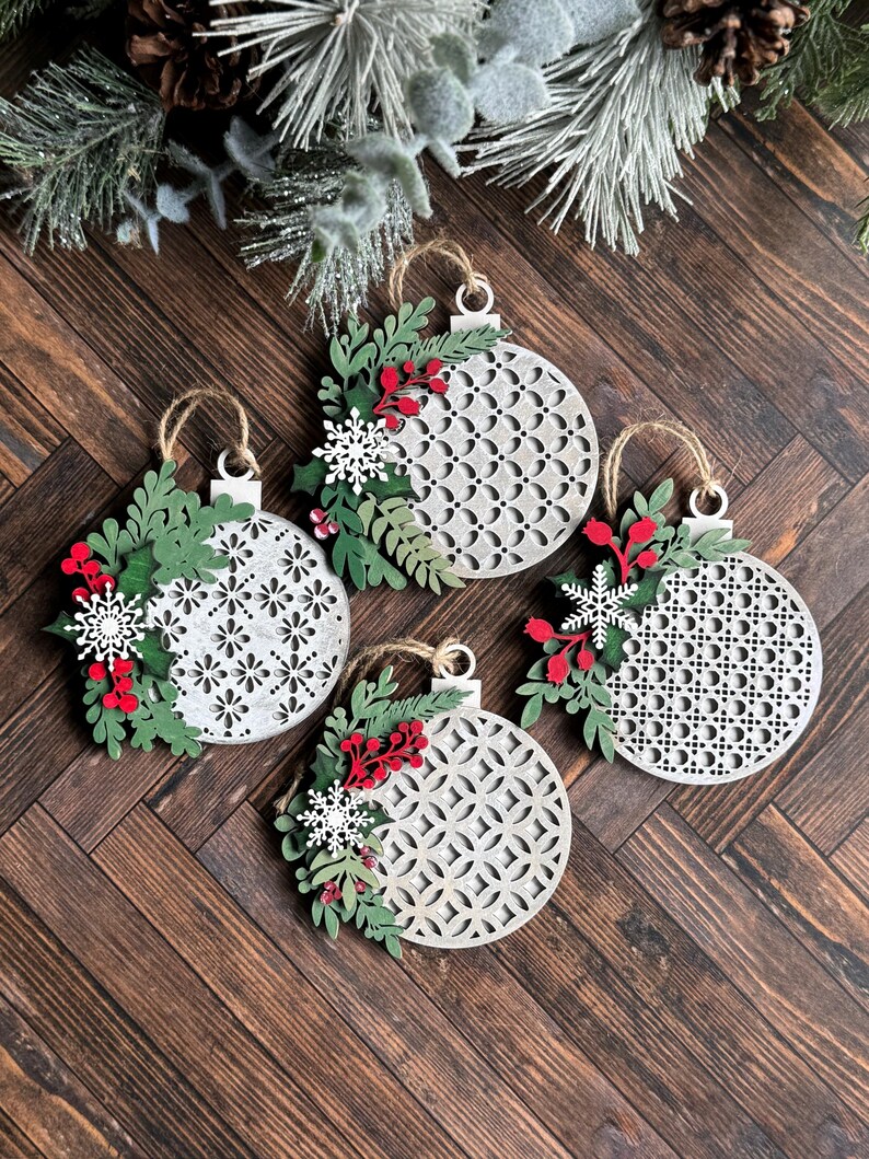 May include: Four white wooden Christmas ornaments with a geometric pattern. Each ornament is decorated with green leaves, red berries, and white snowflakes.