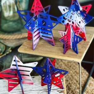 May include: Five wooden stars of varying sizes, painted in a red, white and blue gradient. The stars have a cut-out design and are arranged on a wooden surface.