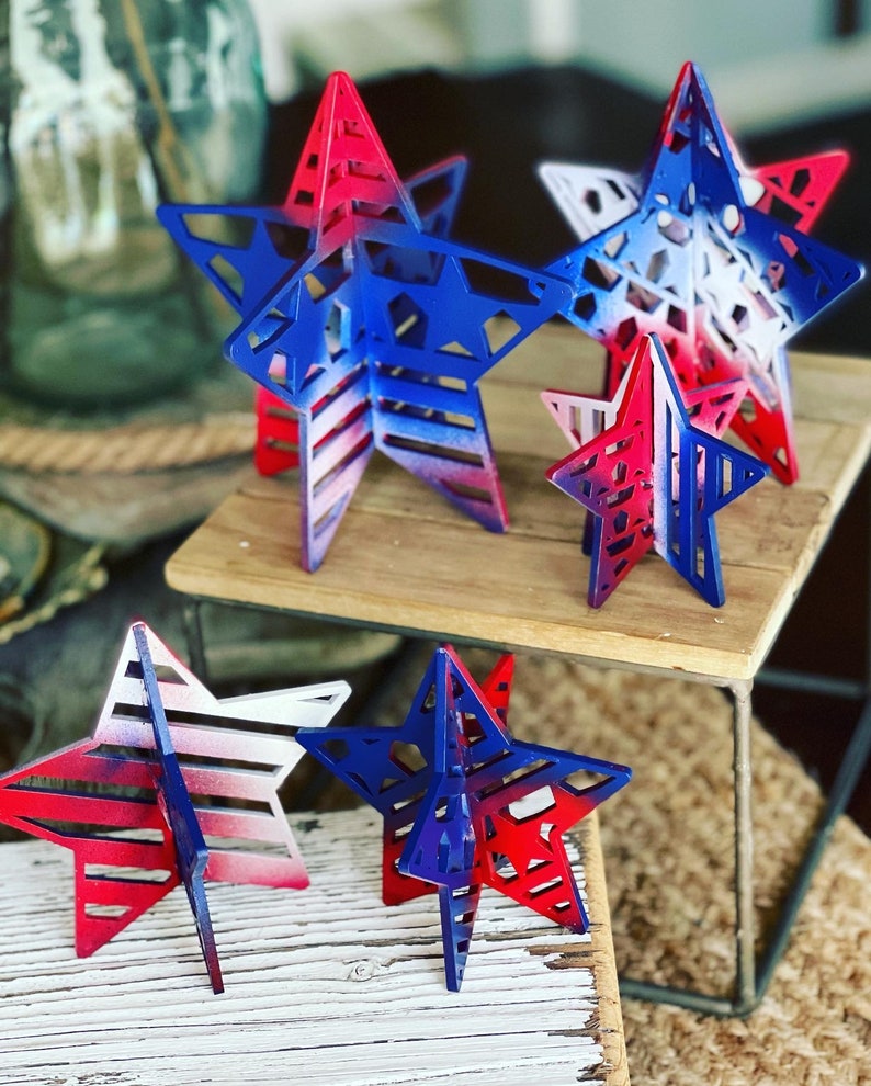 May include: Five wooden stars of varying sizes, painted in a red, white and blue gradient. The stars have a cut-out design and are arranged on a wooden surface.