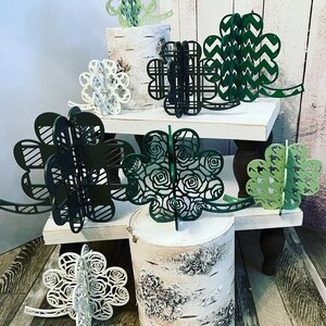 May include: Assortment of green, black, and white paper shamrock cutouts. The shamrocks are layered and have different designs. They are displayed on white wooden stands.