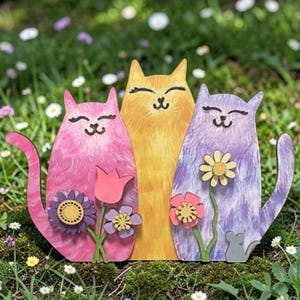 May include: A wooden garden decoration featuring three stylized cats in pink, yellow, and purple. Each cat has a smiling face and is accompanied by a flower. The cats are set against a backdrop of green grass and small white flowers.
