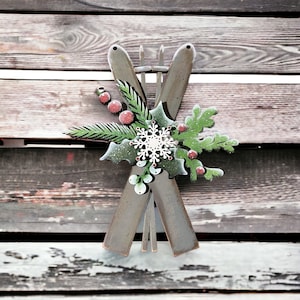May include: Wooden Christmas decoration featuring crossed skis with a festive arrangement of green leaves, red berries, and a white snowflake. The skis are painted in a muted gray color, creating a rustic holiday aesthetic.