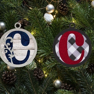 May include: Two Christmas ornaments with the letter "C" are displayed against a backdrop of evergreen branches, pinecones, and silver ornaments. One ornament features a navy blue "C" on a wood-grain background, while the other has a red "C" on a black and white plaid pattern.