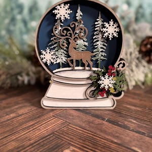 Puede incluir: Un globo de nieve navide&ntilde;o de madera con un ciervo, &aacute;rboles y copos de nieve. El globo tiene un fondo azul oscuro y est&aacute; rodeado por una base blanca. Elementos decorativos incluyen bayas rojas y hojas verdes.