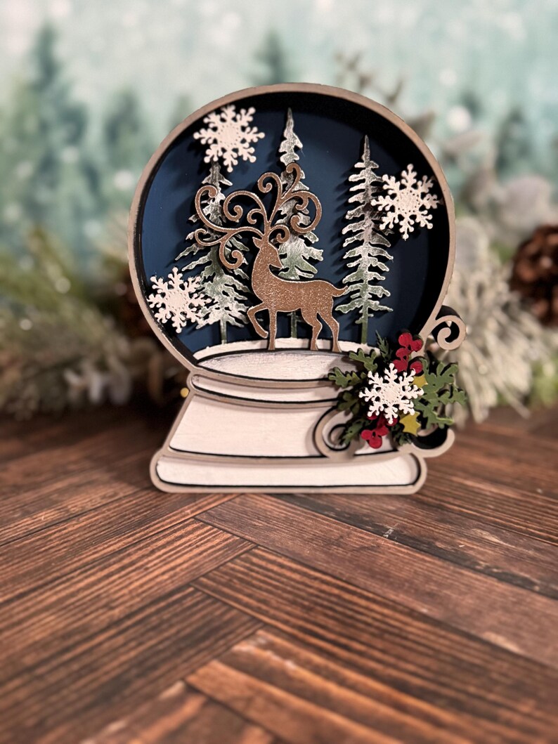 Puede incluir: Un globo de nieve navide&ntilde;o de madera con un ciervo, &aacute;rboles y copos de nieve. El globo tiene un fondo azul oscuro y est&aacute; rodeado por una base blanca. Elementos decorativos incluyen bayas rojas y hojas verdes.