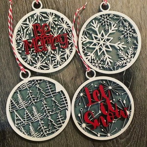 Puede incluir: Cuatro adornos navideños de madera blanca con letras rojas. Los adornos presentan diferentes diseños de temática invernal, incluyendo copos de nieve, árboles y las frases "Sé alegre" y "Deja que nieve".