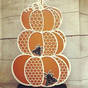 May include: A three-tiered pumpkin decoration with a honeycomb pattern and two black bees. The pumpkin is made of wood and is painted orange. The bees are cut out of black paper and are attached to the pumpkin.
