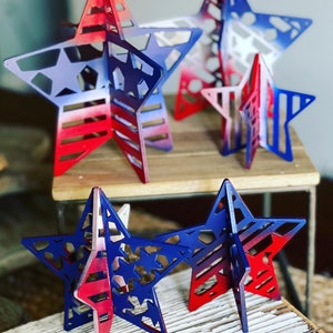 May include: Four wooden stars painted in red, white and blue with a patriotic theme. The stars are layered and have a 3D effect.