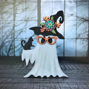 May include: A whimsical Halloween decoration featuring a ghost wearing orange glasses and a black witch hat adorned with colorful flowers. A black cat sits on the ghost's shoulder. The ghost is painted white with gray shading, creating a charming, festive design.