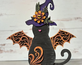 May include: A black cat wearing a purple witch hat with a floral design and orange and yellow accents. The cat has bat wings with a brown and gold filigree design and a small pink heart on its chest.