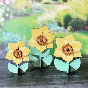 May include: Three wooden daffodil flower decorations with yellow petals and brown centers. The flowers are on green stems with leaves.