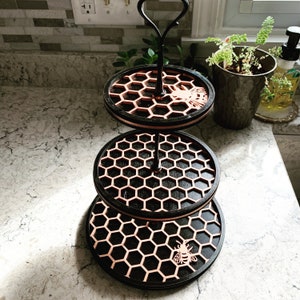 May include: A three-tiered serving tray with a black frame and copper-colored honeycomb design. Each tier features a laser-cut bee. The tray has a black handle and is suitable for displaying desserts or small items.
