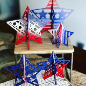 May include: Five wooden stars of varying sizes, painted in a red, white and blue gradient. The stars are standing on a wooden surface.