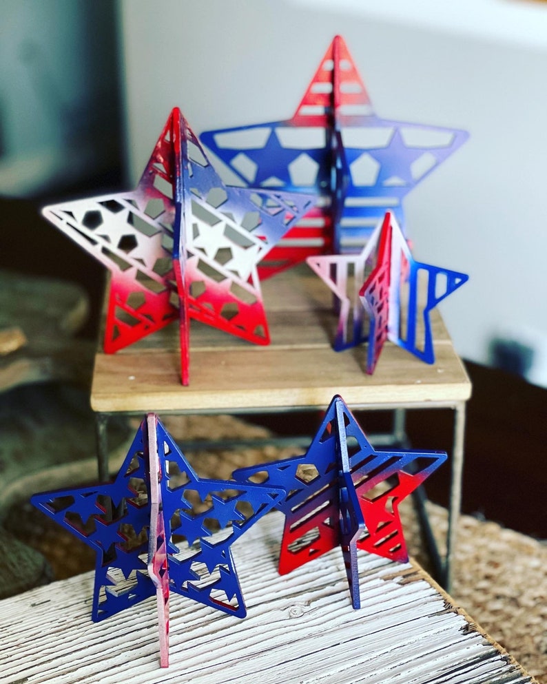 May include: Five wooden stars of varying sizes, painted in a red, white and blue gradient. The stars are standing on a wooden surface.