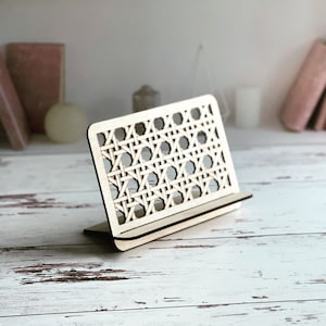 May include: A wooden phone stand with a woven pattern. The stand is made of light brown wood and has a small shelf for holding a phone.