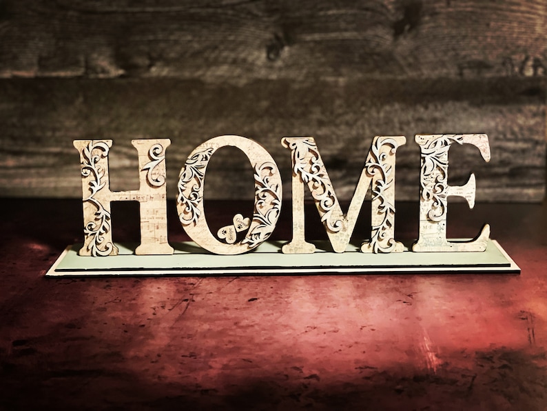 Może przedstawiać: Drewniany znak z napisem "HOME" z misternymi, kwiatowymi wycięciami. Beżowe litery mają postarzaną fakturę i są umieszczone na prostokątnej podstawie. Tło to ciemna drewniana powierzchnia, idealna do dekoracji domu.