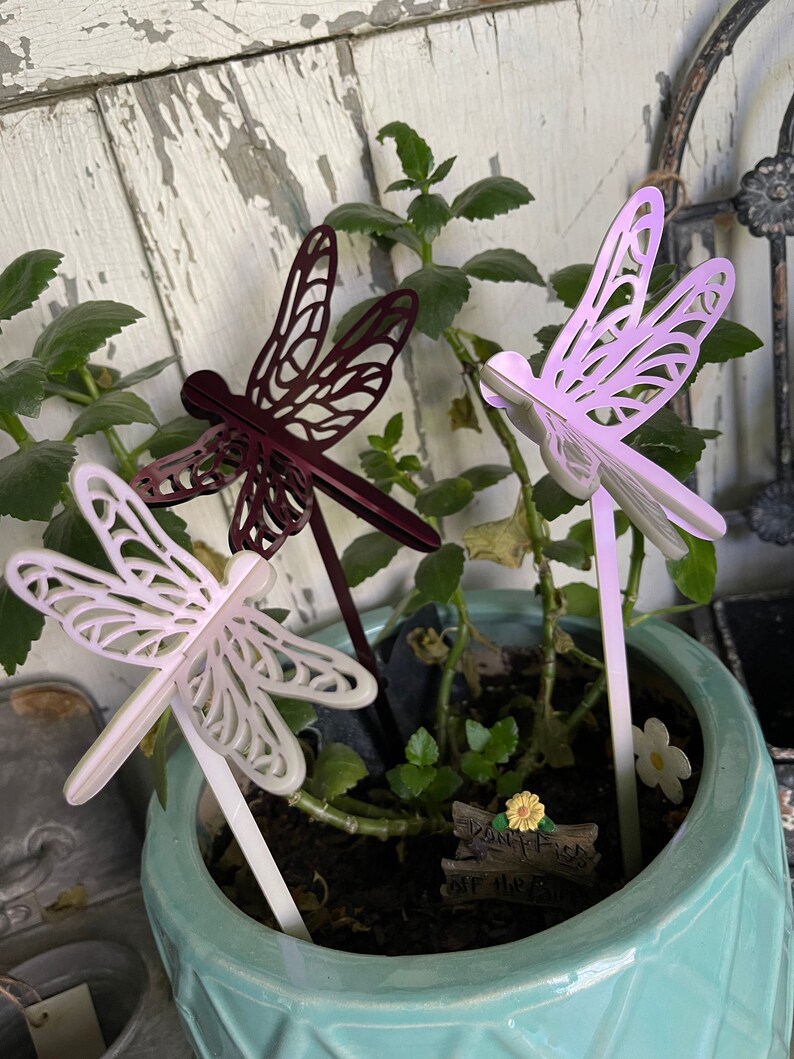 May include: Three decorative metal dragonfly garden stakes in a pot of green plants. The dragonflies are white, purple, and maroon.