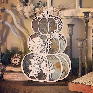 May include: A decorative metal pumpkin stack with a distressed finish and intricate floral cutouts. The pumpkins are stacked on a black metal base.