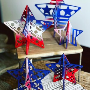 May include: Four wooden stars painted in red, white and blue with a distressed paint finish. The stars are different sizes and are standing on a wooden surface.