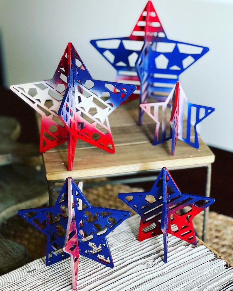 May include: Four wooden stars painted in red, white and blue with a distressed paint finish. The stars are different sizes and are standing on a wooden surface.