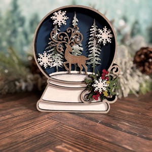 Puede incluir: Decoraci&oacute;n de globo de nieve de madera con un ciervo, &aacute;rboles y copos de nieve. El globo tiene un fondo azul oscuro y una base blanca. Se adjuntan vegetaci&oacute;n decorativa y bayas rojas a la base. El tema es invierno y Navidad.