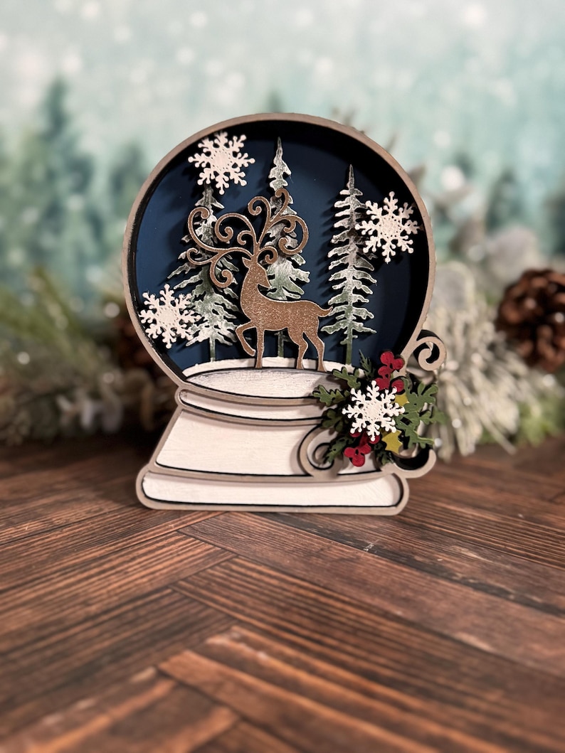Puede incluir: Decoraci&oacute;n de globo de nieve de madera con un ciervo, &aacute;rboles y copos de nieve. El globo tiene un fondo azul oscuro y una base blanca. Se adjuntan vegetaci&oacute;n decorativa y bayas rojas a la base. El tema es invierno y Navidad.