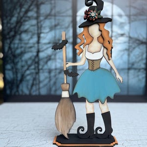 May include: Wooden Halloween witch figurine with a black hat adorned with flowers, orange hair, and a blue skirt. The witch holds a broom with bat accents. The figurine stands on a black base with an orange trim.