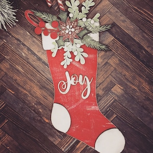 Puede incluir: Calcetín de Navidad rojo con ribete blanco y la palabra "Alegría" escrita en blanco. El calcetín está decorado con un arreglo floral verde y blanco y un bastón de caramelo.