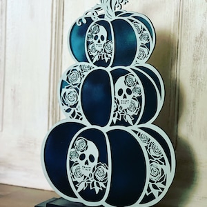 Może przedstawiać: Trójwarstwowa drewniana dekoracja w kształcie dyni z ciemnoniebieskim wykończeniem. Każda dynia jest misterne rzeźbiona z białymi czaszkami i różami.