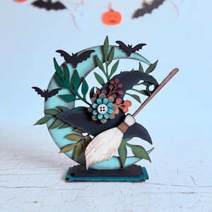 Puede incluir: Decoración de madera con temática de Halloween. Presenta una luna creciente turquesa, un sombrero de bruja y una escoba. El sombrero está adornado con flores y un botón. También se incluyen murciélagos y hojas. La paleta de colores incluye turquesa, negro, marrón y naranja.