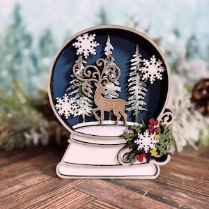 Puede incluir: Globo de nieve decorativo de madera con un ciervo, copos de nieve y pinos. El globo tiene un interior azul oscuro y detalles en blanco, dorado, rojo y verde. La base es blanca con ribete negro, ideal para la decoraci&oacute;n navide&ntilde;a.