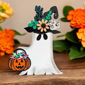 May include: A white ghost wearing a black witch hat with green and orange floral accents. The ghost is holding a small orange pumpkin with colorful candy inside.