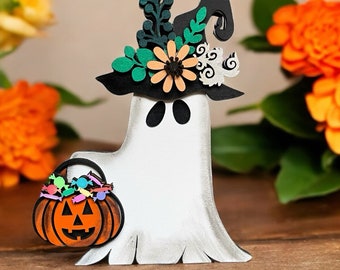 May include: A white ghost wearing a black witch hat with green and orange floral accents. The ghost is holding a small orange pumpkin with colorful candy inside.