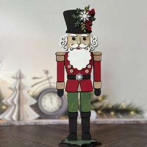 May include: A wooden Christmas nutcracker figurine. The figurine has a black top hat, a white beard, and a red jacket with gold buttons. It is decorated with a snowflake and greenery on the hat and a snowflake on the jacket.