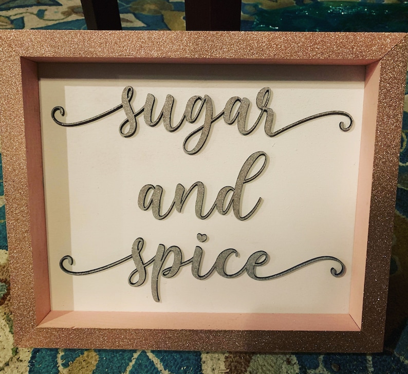 Puede incluir: Un letrero enmarcado con purpurina rosa con las palabras "sugar and spice" escritas en cursiva sobre un fondo blanco.