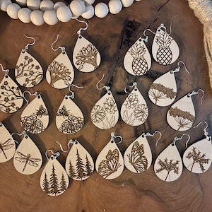 May include: A collection of 16 pairs of teardrop-shaped wooden earrings with various laser-cut designs, including butterflies, dragonflies, flowers, trees, and pineapples. The earrings are all a light brown color and have silver hooks.