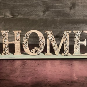 Może przedstawiać: Dekoracyjny drewniany znak z napisem "HOME". Jasne litery mają skomplikowane kwiatowe wycięcia i wz&oacute;r vintage papieru. Znak stoi na jasnoniebieskiej podstawie, idealny do dekoracji domu.