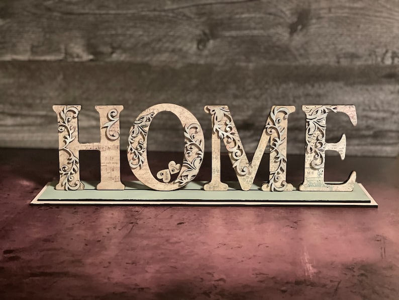 Może przedstawiać: Dekoracyjny drewniany znak z napisem "HOME". Jasne litery mają skomplikowane kwiatowe wycięcia i wz&oacute;r vintage papieru. Znak stoi na jasnoniebieskiej podstawie, idealny do dekoracji domu.