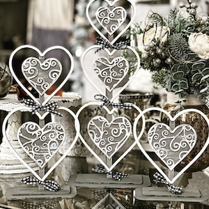 Puede incluir: Conjunto de cinco decoraciones de metal blanco en forma de corazón con un diseño de filigrana gris. Los corazones están dispuestos en una pila escalonada, con el corazón más grande en la parte inferior y el corazón más pequeño en la parte superior. Cada corazón está sobre una base gris.
