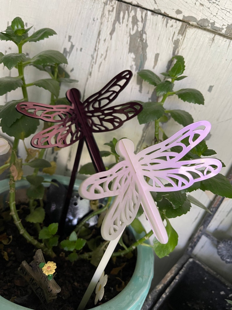 May include: Two decorative metal dragonfly garden stakes. One is purple and the other is white. The stakes are inserted into a pot of green plants.