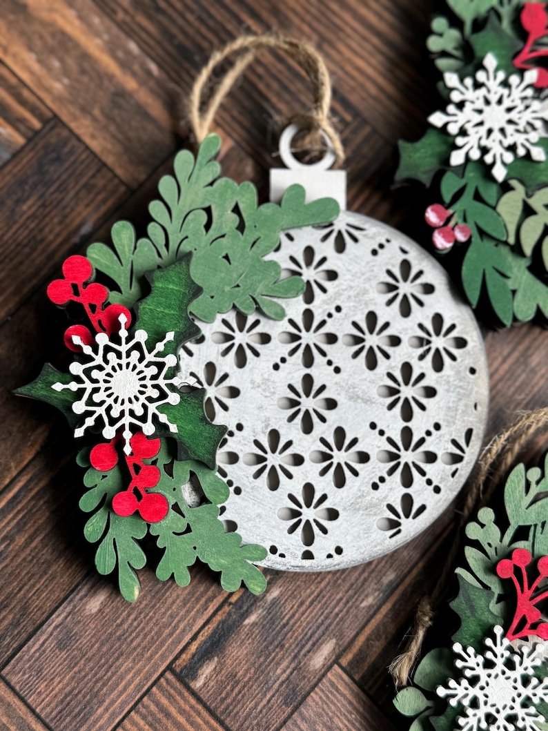 May include: A white, wooden, decorative Christmas ornament with a cut-out design. The ornament is decorated with green and red paper leaves and a white snowflake.