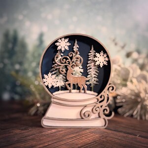 Puede incluir: Globo de nieve decorativo de madera con un ciervo, &aacute;rboles y copos de nieve. El globo tiene un fondo azul oscuro y est&aacute; sobre una base de madera. El dise&ntilde;o es intrincado y detallado, con un tema invernal.