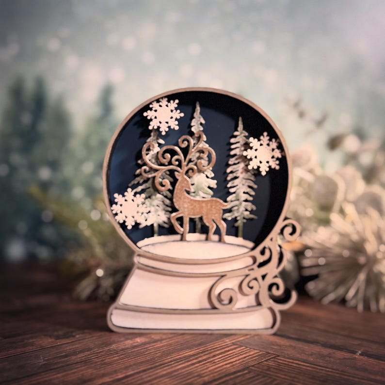 Puede incluir: Globo de nieve decorativo de madera con un ciervo, &aacute;rboles y copos de nieve. El globo tiene un fondo azul oscuro y est&aacute; sobre una base de madera. El dise&ntilde;o es intrincado y detallado, con un tema invernal.