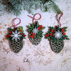 May include: Three festive Christmas tree ornaments. Each is shaped like a pine cone and decorated with green leaves, red berries, and a white snowflake. They hang from red and white twine. The background has a patterned design.